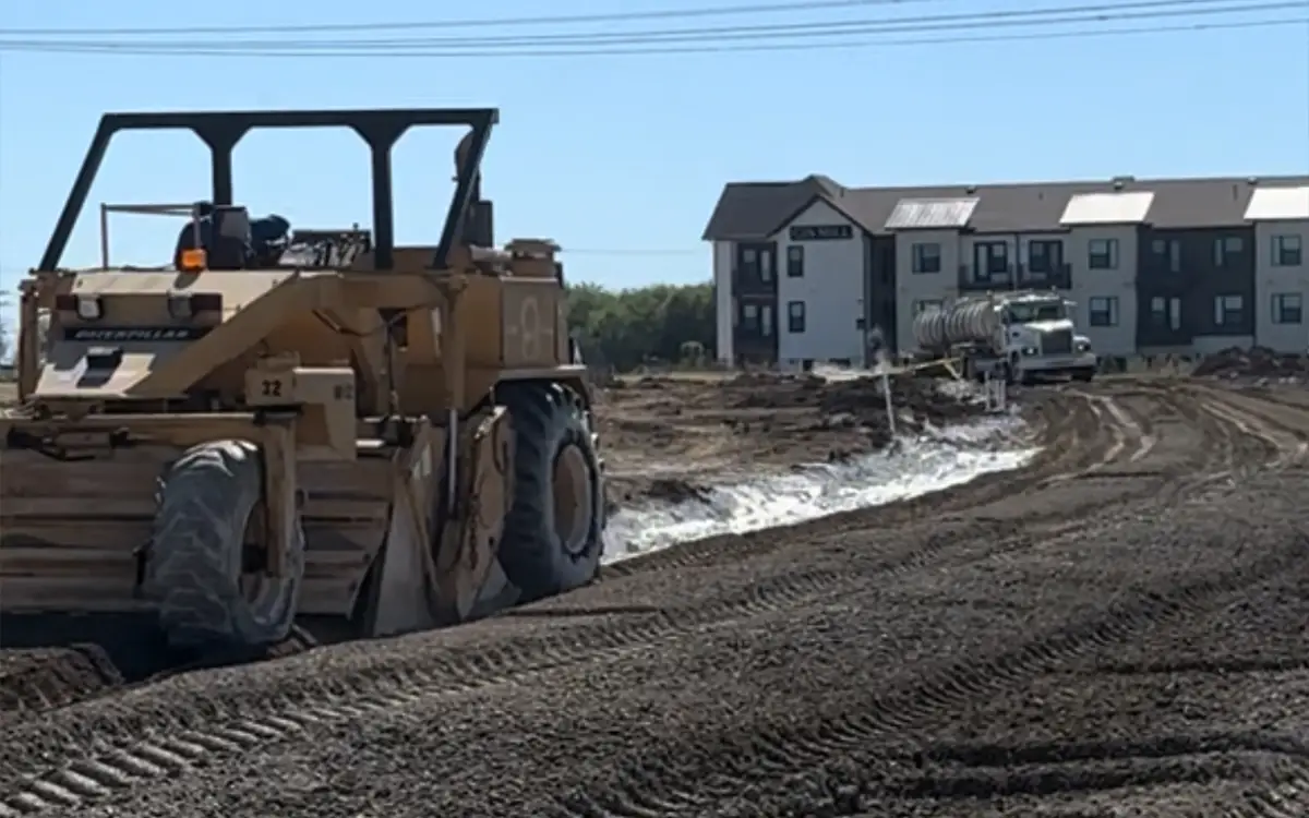 dirt with snow on it with a machine working on stomping the dirt with a house with snow on top of the roof behind it