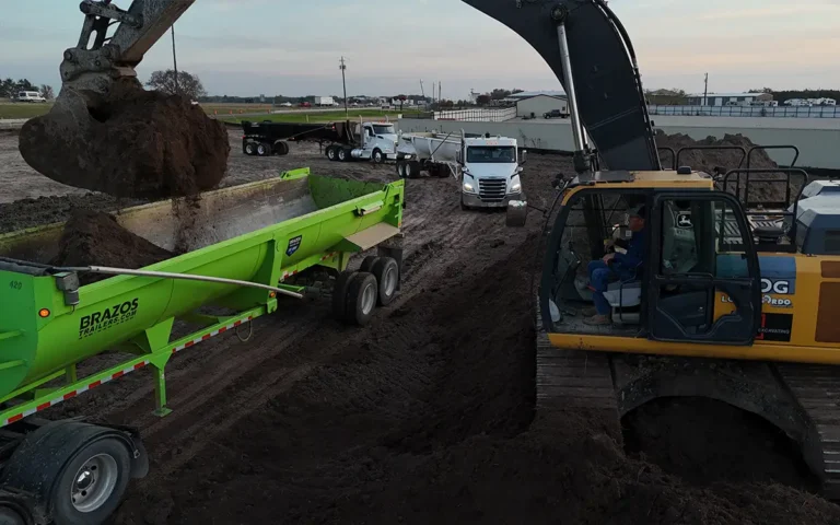 Excavator loading dirt into a green Brazos Equipment trailer at a construction site with trucks in the background.