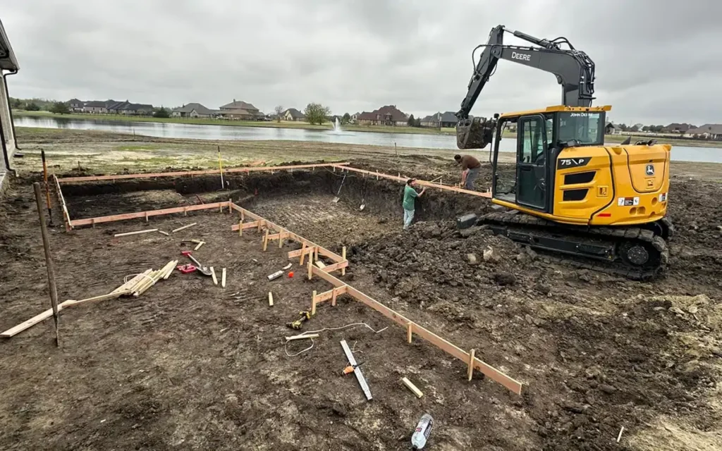 Excavation crew from B Squared Excavating LLC digging a foundation with a John Deere excavator next to a pond.